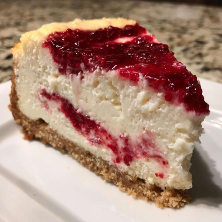 A thick slice of creamy white chocolate cheesecake featuring a bright red raspberry swirl and topping, resting on a graham cracker crust.