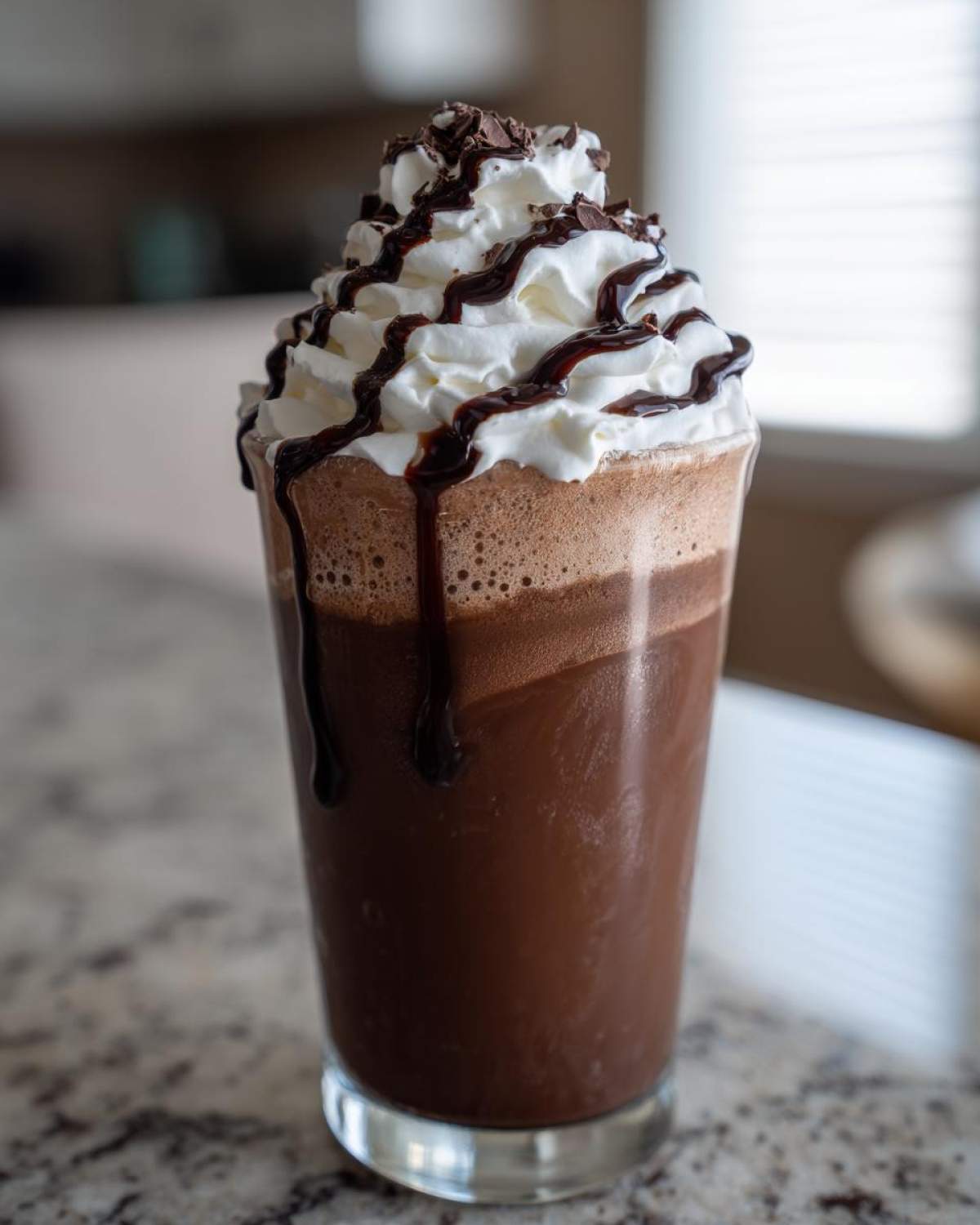 A tall glass filled with rich, blended frozen hot chocolate, topped with whipped cream, chocolate syrup drizzle, and shavings.