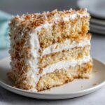 A tall, moist slice of layered coconut cake with white frosting and toasted coconut flakes.