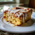 A close-up of a square serving of fluffy cinnamon roll casserole topped with thick white icing dripping down the sides.
