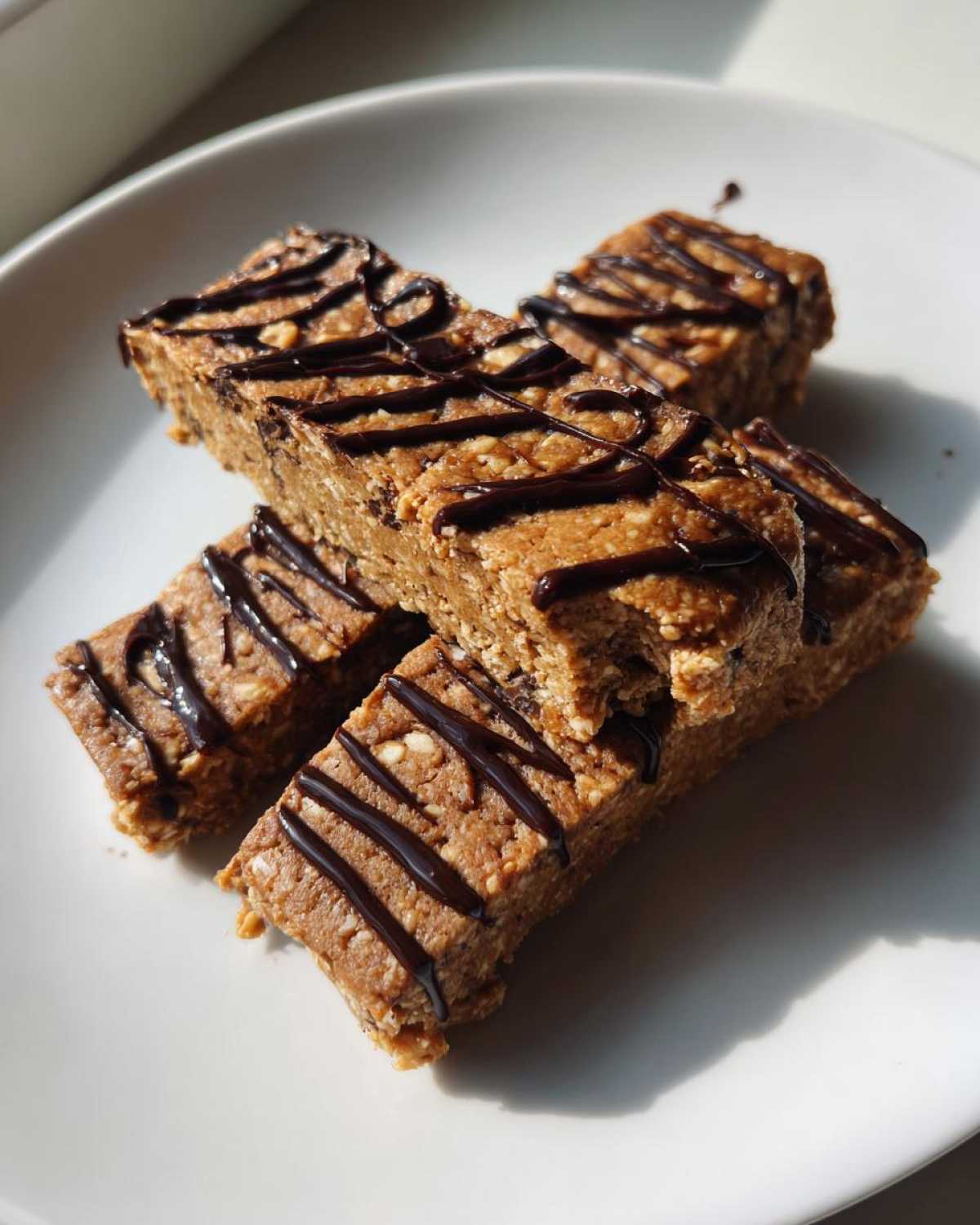 Four homemade protein bars stacked on a white plate, drizzled with dark chocolate.