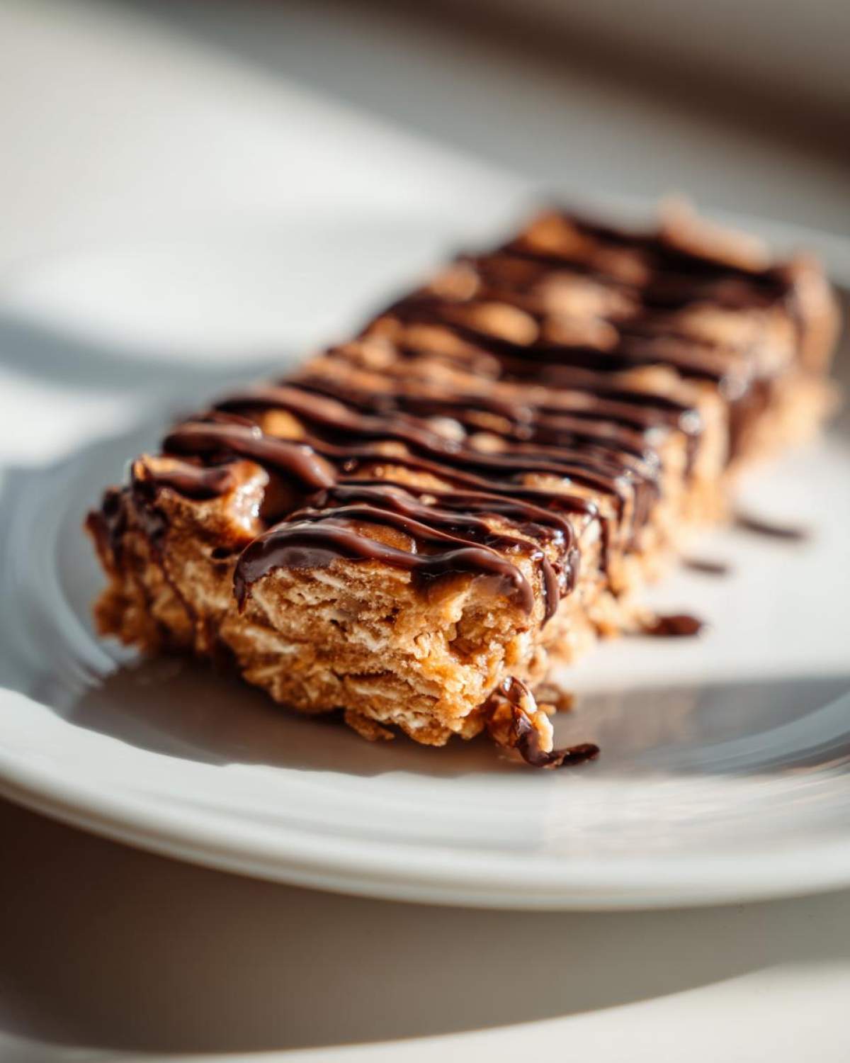 Close-up of a homemade, chewy protein bar drizzled with dark chocolate, sitting on a white plate.