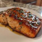 A close-up of a beautifully glazed and cooked piece of air fryer salmon, topped with herbs, resting on a white plate.