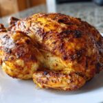 A whole roasted chicken with crispy, seasoned skin cooked in an air fryer whole chicken recipe, resting on a white plate.