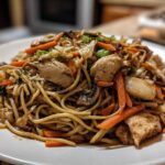 A close-up of a generous serving of chicken chow mein recipe featuring noodles, sliced chicken, carrots, and mushrooms on a white plate.