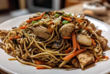 A close-up of a generous serving of chicken chow mein recipe featuring noodles, sliced chicken, carrots, and mushrooms on a white plate.