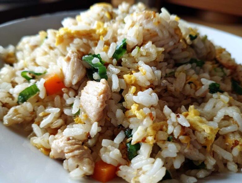 Close-up macro shot of fluffy white rice mixed with chunks of chicken, scrambled egg, carrots, and green onions in chicken fried rice.