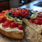 A slice removed from a fruit pizza sugar cookie topped with cream cheese frosting, strawberries, blueberries, and kiwi.