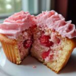 Close-up of a strawberry cupcake cut in half showing real strawberry pieces inside the cake and topped with pink strawberry frosting.