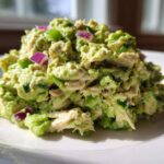 Close-up of a mound of creamy avocado chicken salad featuring shredded chicken, green celery, and diced red onion on a white plate.