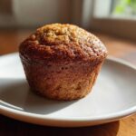 A single, perfectly baked banana protein muffin with a golden-brown, slightly glossy top, resting on a small white plate.