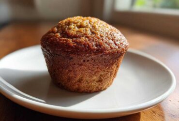 A single, perfectly baked banana protein muffin with a golden-brown, slightly glossy top, resting on a small white plate.