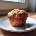 A single, perfectly baked banana protein muffin sitting on a small white plate, backlit by window light.