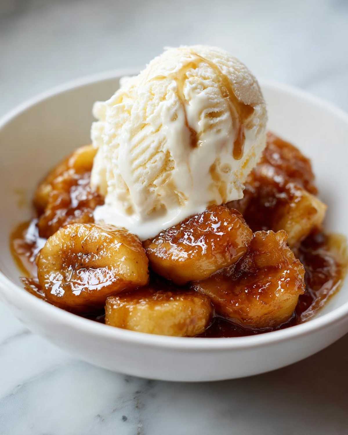 A white bowl filled with caramelized bananas in sauce, topped with a scoop of melting vanilla ice cream, ready to eat Bananas Foster.