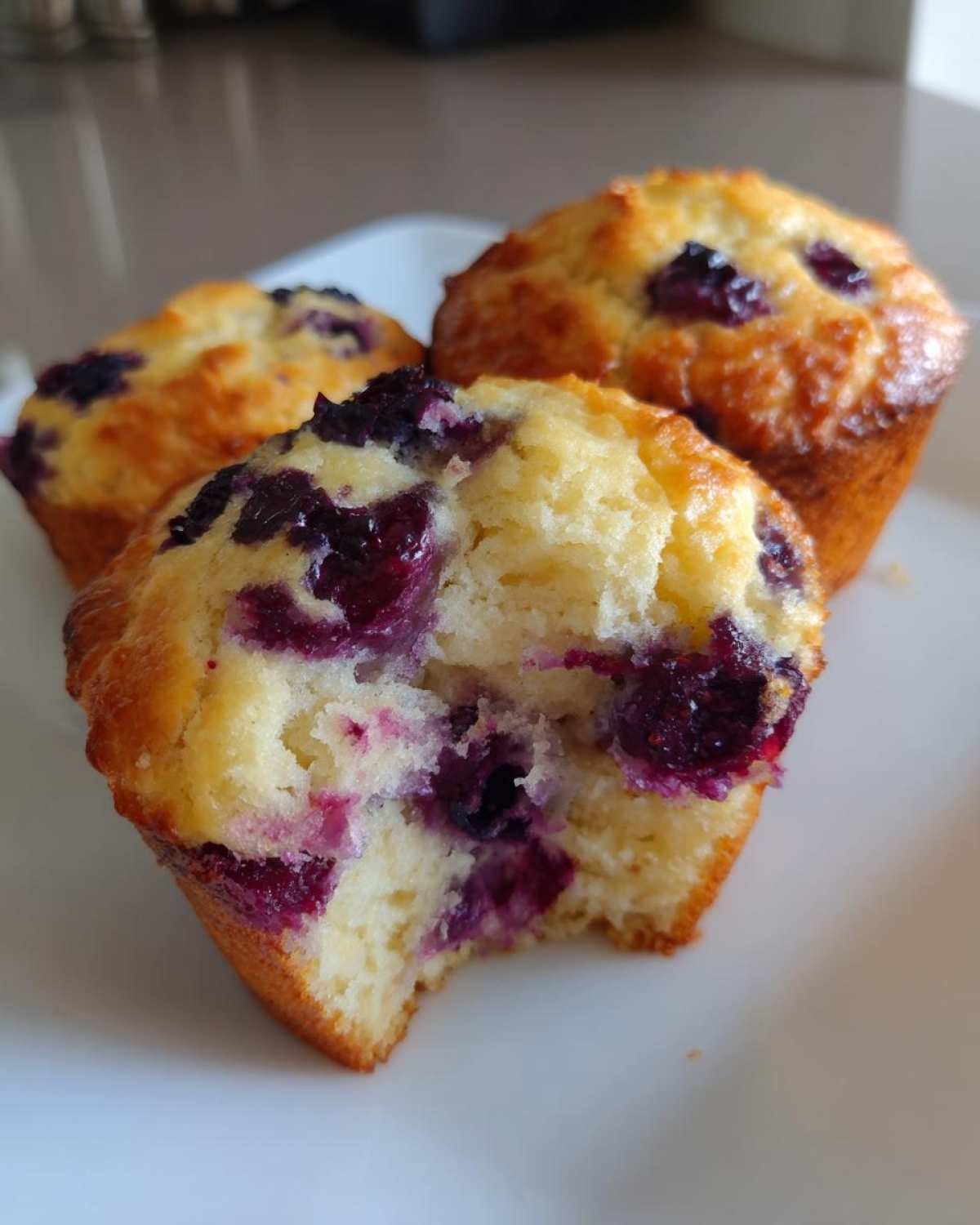 Close-up of three blueberry protein muffins on a white plate, one of which has a bite taken out showing the moist interior.