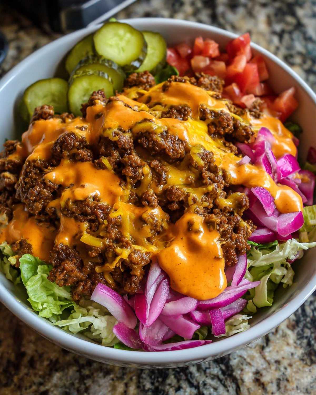 A close-up view of a delicious cheeseburger bowl featuring seasoned ground beef, melted cheese, special sauce, lettuce, red onion, pickles, and tomatoes.