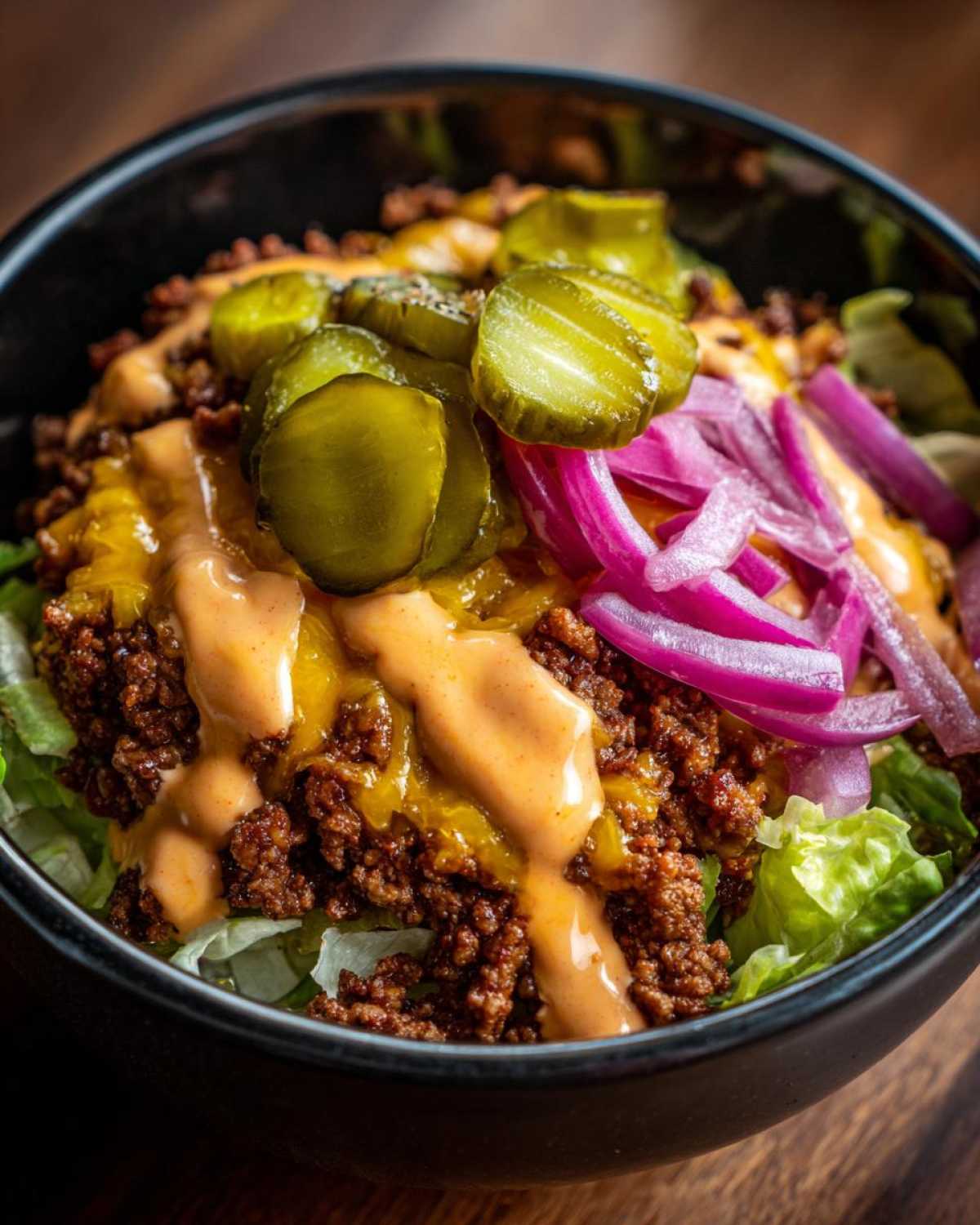 Close-up of a cheeseburger bowl featuring seasoned ground beef, melted cheese, special sauce, pickles, and red onions over lettuce.