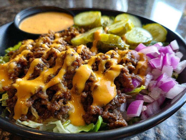 A close-up of a delicious cheeseburger bowl featuring seasoned ground beef, melted cheese sauce, diced red onion, and pickles.