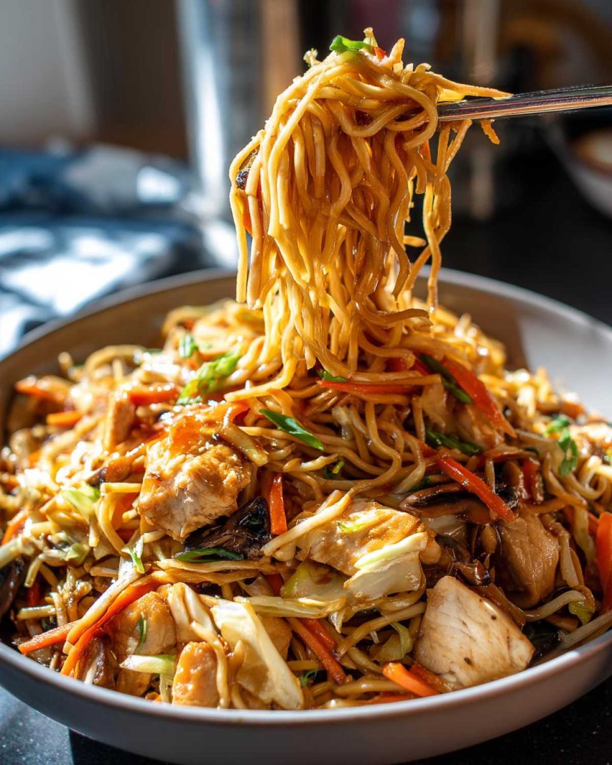 Chopsticks lifting a large portion of noodles from a bowl of savory chicken chow mein recipe with vegetables.