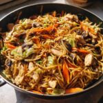 Close-up of a skillet filled with freshly made chicken chow mein recipe, featuring noodles, chicken pieces, carrots, and cabbage.