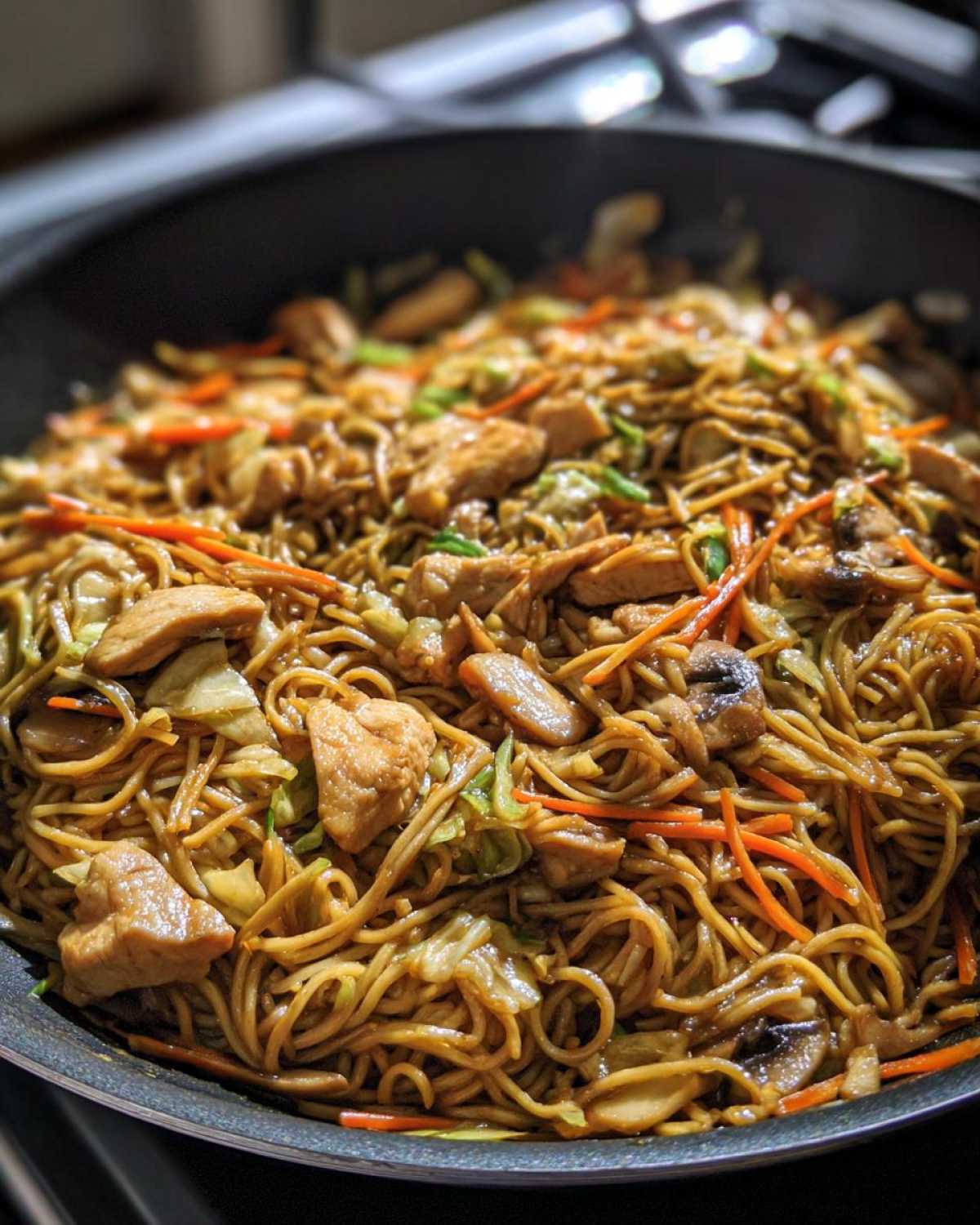 Close-up of delicious chicken chow mein recipe being cooked in a large dark wok with noodles, chicken pieces, and vegetables.