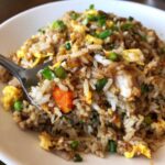 A spoonful of savory chicken fried rice showing rice, chicken pieces, scrambled egg, carrots, and green onions.
