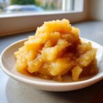 Close-up of chunky, golden homemade applesauce served in a small white bowl near a window.