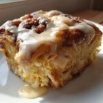 A close-up of a square serving of moist Cinnamon roll casserole topped with thick, white icing dripping down the side.