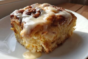 A close-up of a square serving of moist Cinnamon roll casserole topped with thick, white icing dripping down the side.