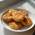 A stack of freshly baked peanut butter cookies featuring the classic criss-cross fork marks, served on a white plate.