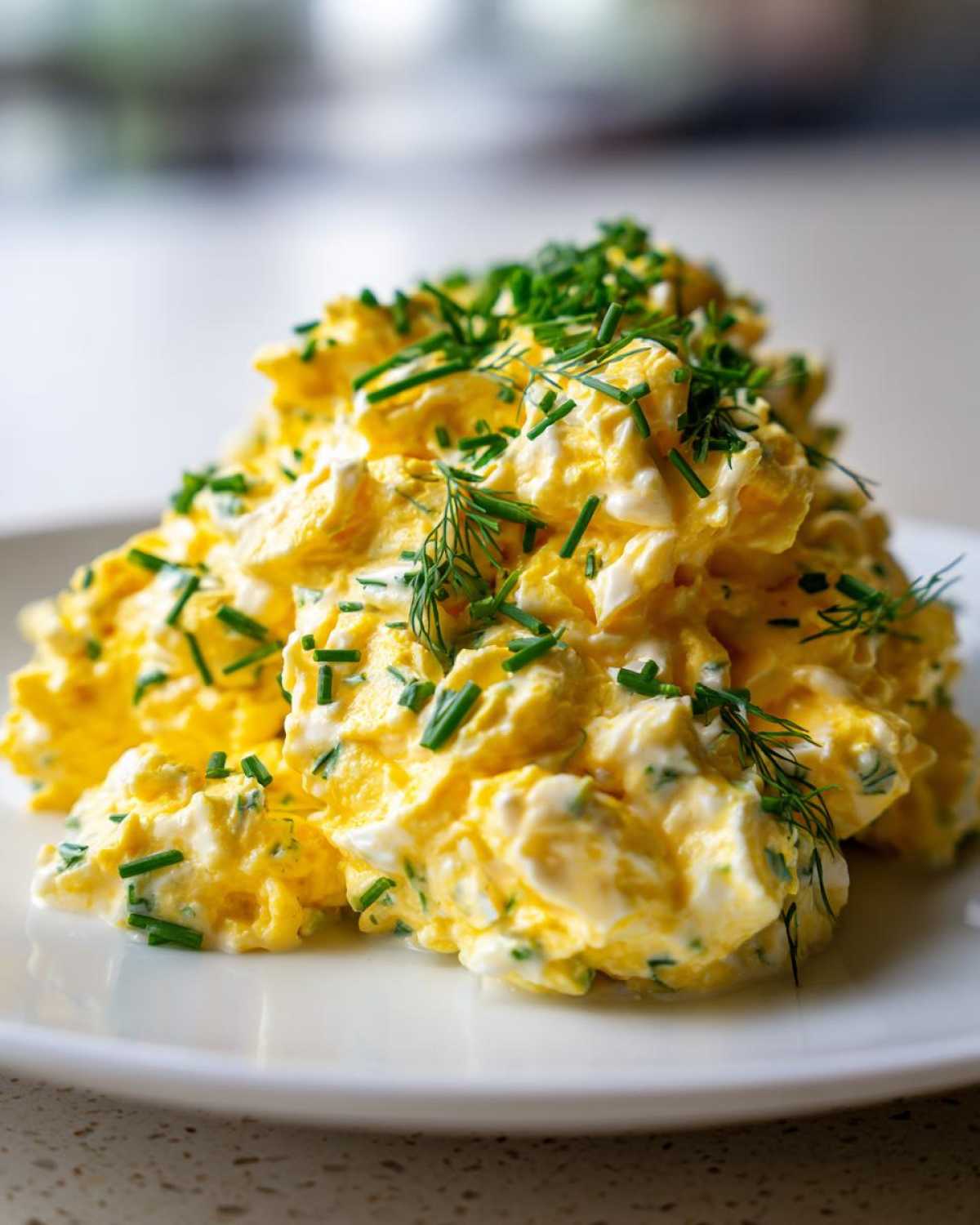 A generous mound of creamy, yellow egg salad recipe garnished with fresh chopped chives and dill.