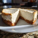 Close-up of a small, round Protein Cheesecake with a thick graham cracker crust, sliced open on a white plate.