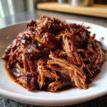 Close-up of tender, shredded crock pot pulled pork coated in a thick, dark BBQ sauce, served on a white plate.