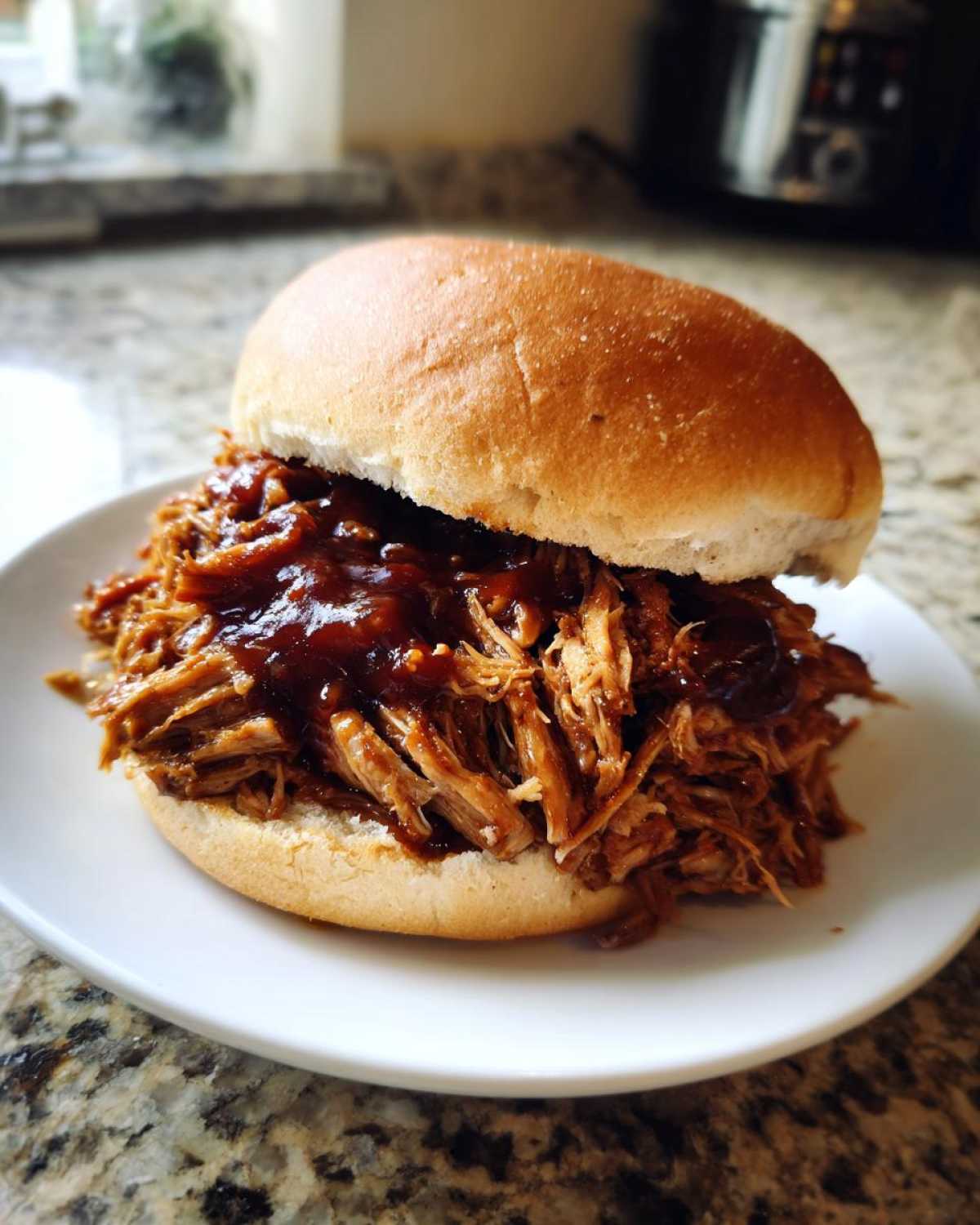 A close-up of a generous serving of saucy crock pot pulled pork piled high on a soft bun, served on a white plate.