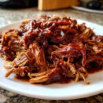Close-up of tender, shredded crock pot pulled pork smothered in rich BBQ sauce on a white plate.