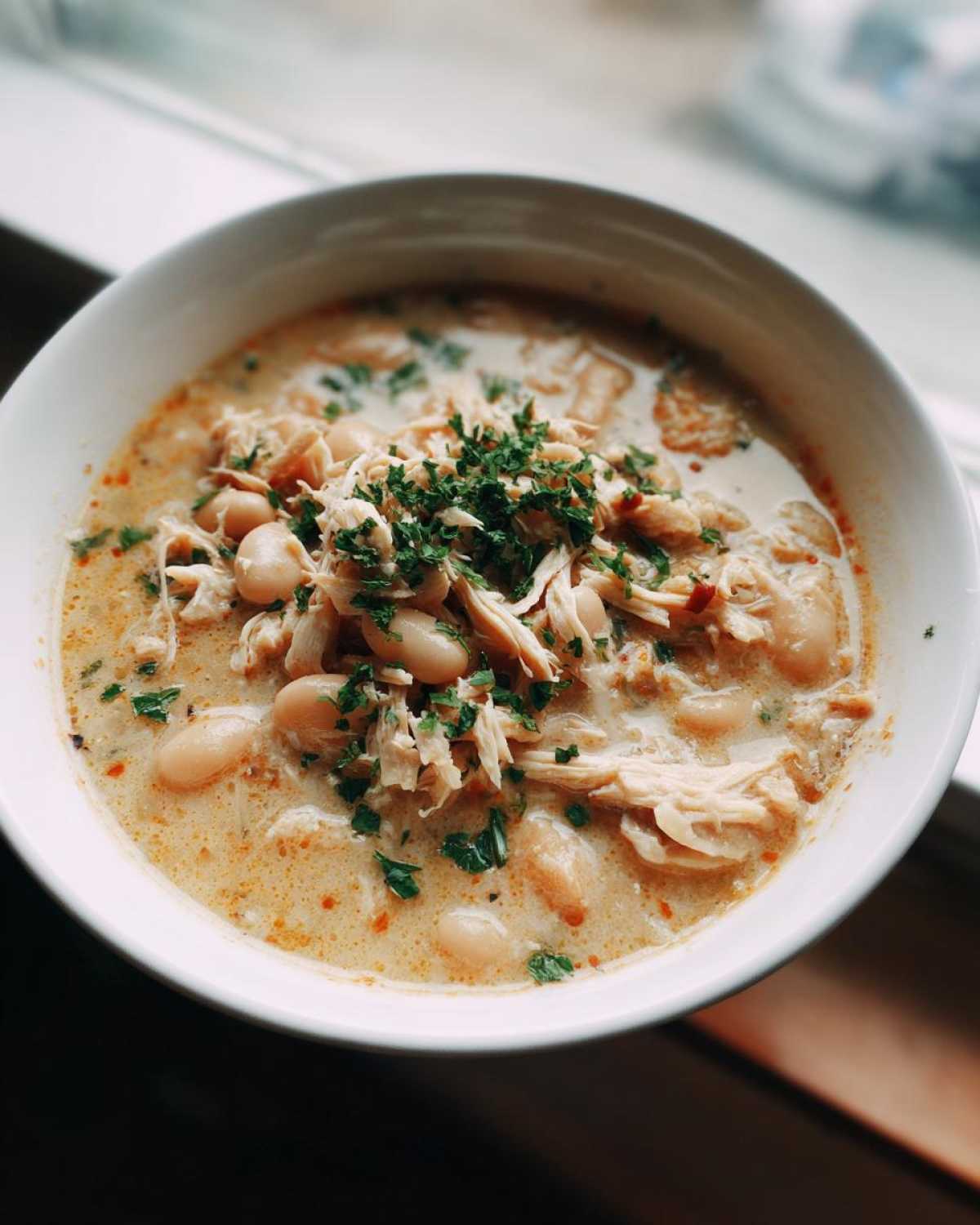 A close-up overhead shot of a white bowl filled with creamy crockpot white chicken chili, topped with shredded chicken, white beans, and fresh parsley.