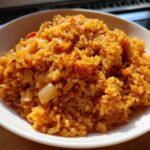 A close-up, brightly lit photo of a bowl filled with flavorful, orange-hued Spanish rice mixed with visible pieces of sautéed onion.
