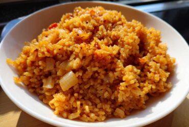 A close-up, brightly lit photo of a bowl filled with flavorful, orange-hued Spanish rice mixed with visible pieces of sautéed onion.