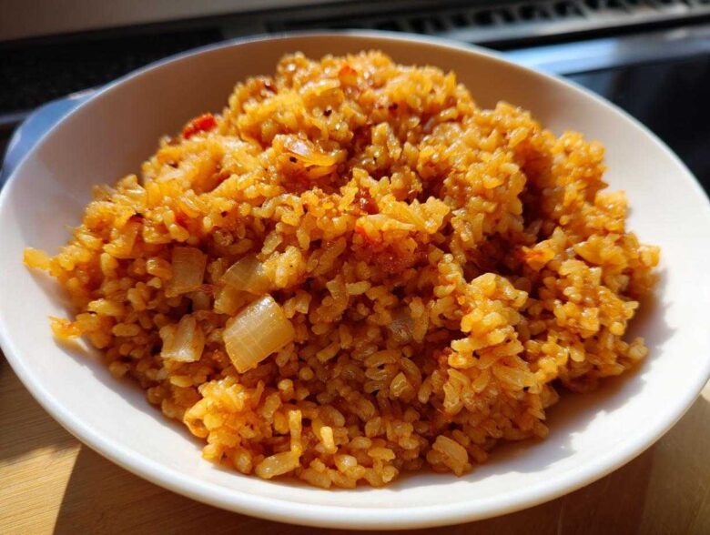 A close-up, brightly lit photo of a bowl filled with flavorful, orange-hued Spanish rice mixed with visible pieces of sautéed onion.