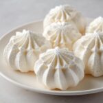 Close-up of several perfectly piped, white meringue cookies with star-shaped tops resting on a white plate.
