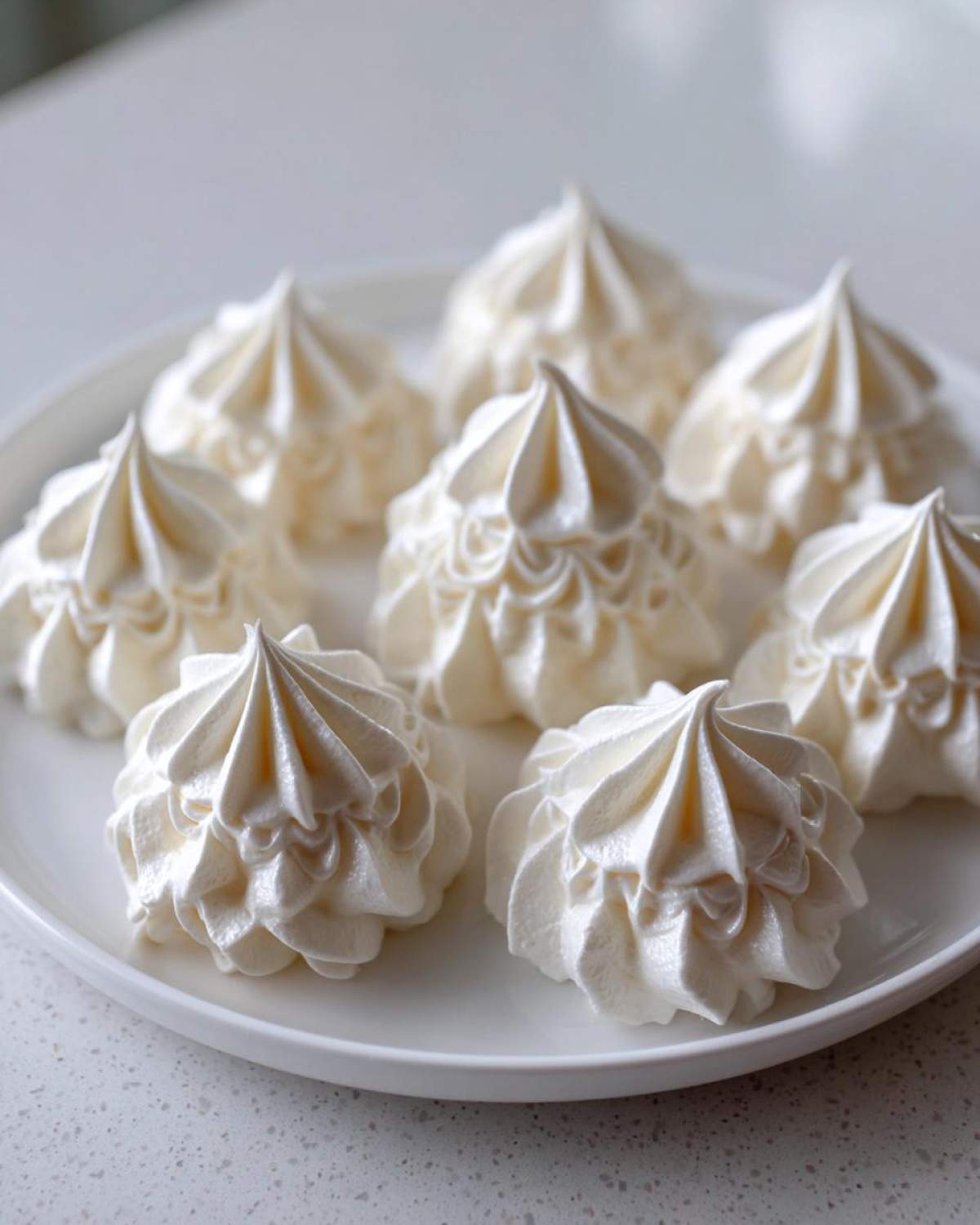 A close-up of several perfectly piped, glossy white meringue cookies arranged on a white serving plate.