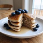 A stack of fluffy almond flour pancakes cut open to show the texture, topped with fresh blueberries and drizzled with syrup.