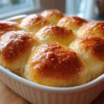 Close-up of golden brown, fluffy dinner rolls baked together in a white dish, glistening with butter glaze.