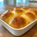 Close-up of golden brown, fluffy dinner rolls baked together in a white ceramic dish, glistening with butter.