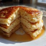 A stack of four fluffy greek yogurt pancakes, cut open to show the texture, drizzled with syrup on a white plate.