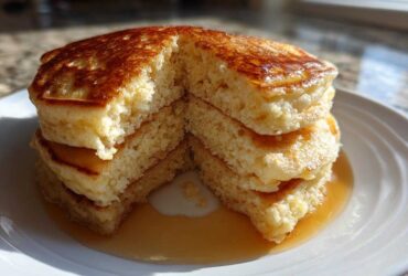 A stack of four fluffy greek yogurt pancakes, cut open to show the texture, drizzled with syrup on a white plate.
