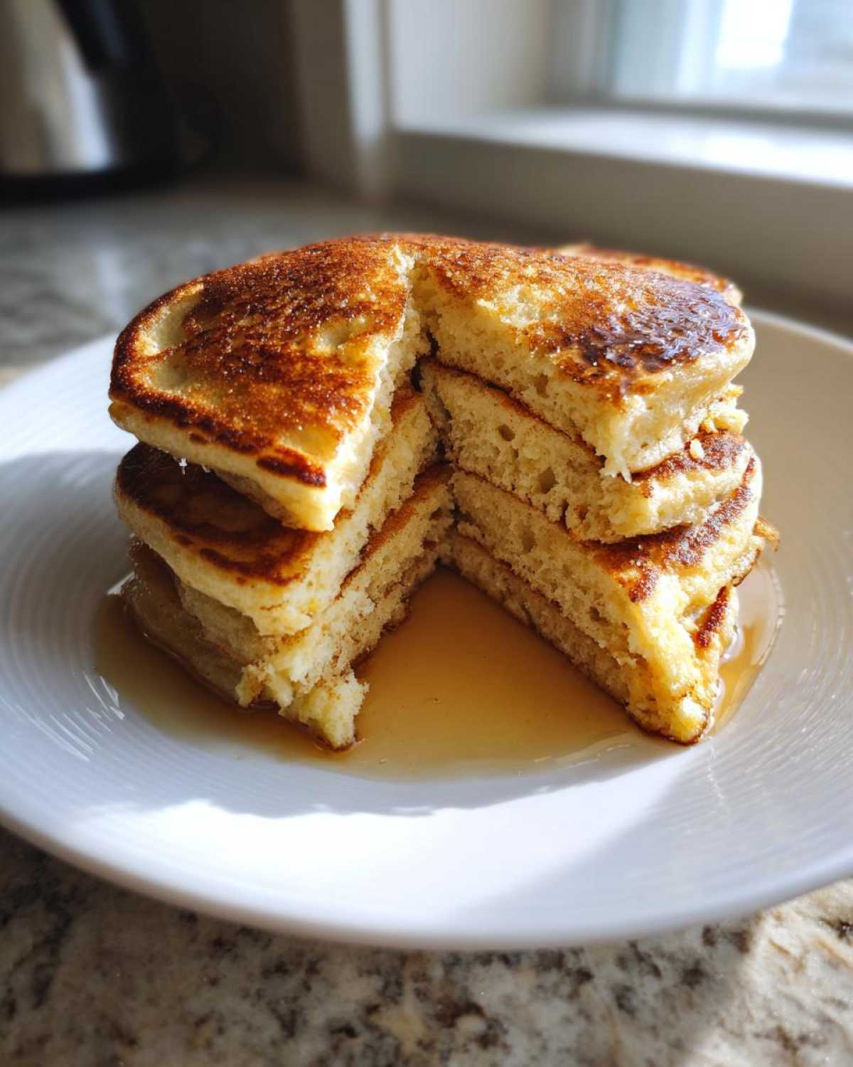 A stack of fluffy greek yogurt pancakes, cut open to show the texture, drizzled with syrup on a white plate.