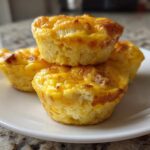 Four fluffy muffin tin egg bites stacked on a white plate, showing a golden-brown, cheesy top.