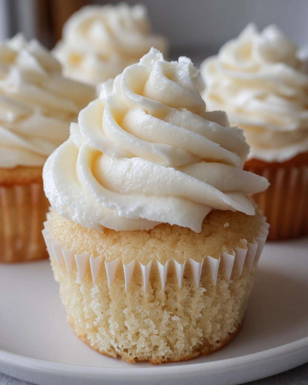 A close-up of one perfectly baked vanilla cupcake topped with a swirl of white buttercream frosting, with more vanilla cupcakes blurred in the background.
