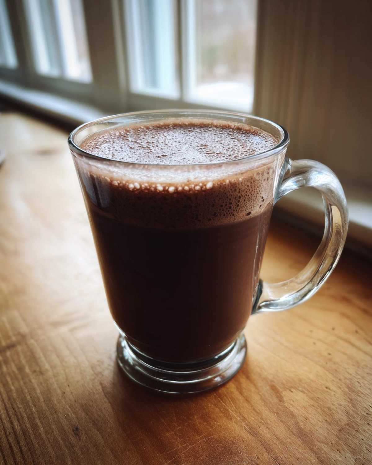 A clear glass mug filled with rich, dark hot cocoa mix topped with foam, sitting on a wooden surface near a window.